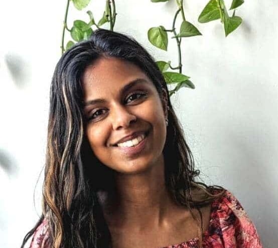 A headshot of a smiling girl wearing a pink and red floral top standing in front of a white grey wall with a green pothos plant hanging behind her